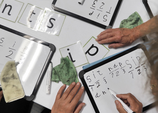 A table covered in letters and minature whiteboards. Two sets of hands can be seen practicing creating words on the whiteboards. 