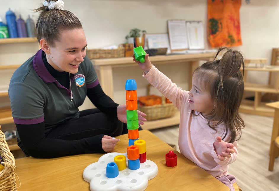A Goodstart Tuggerah educator looks at a female child building a tower. 