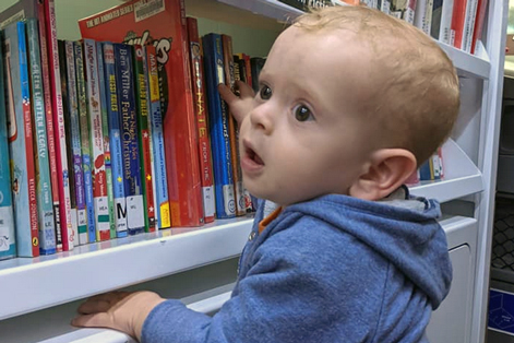 A toddler reaches for a book on the bottom shelf.