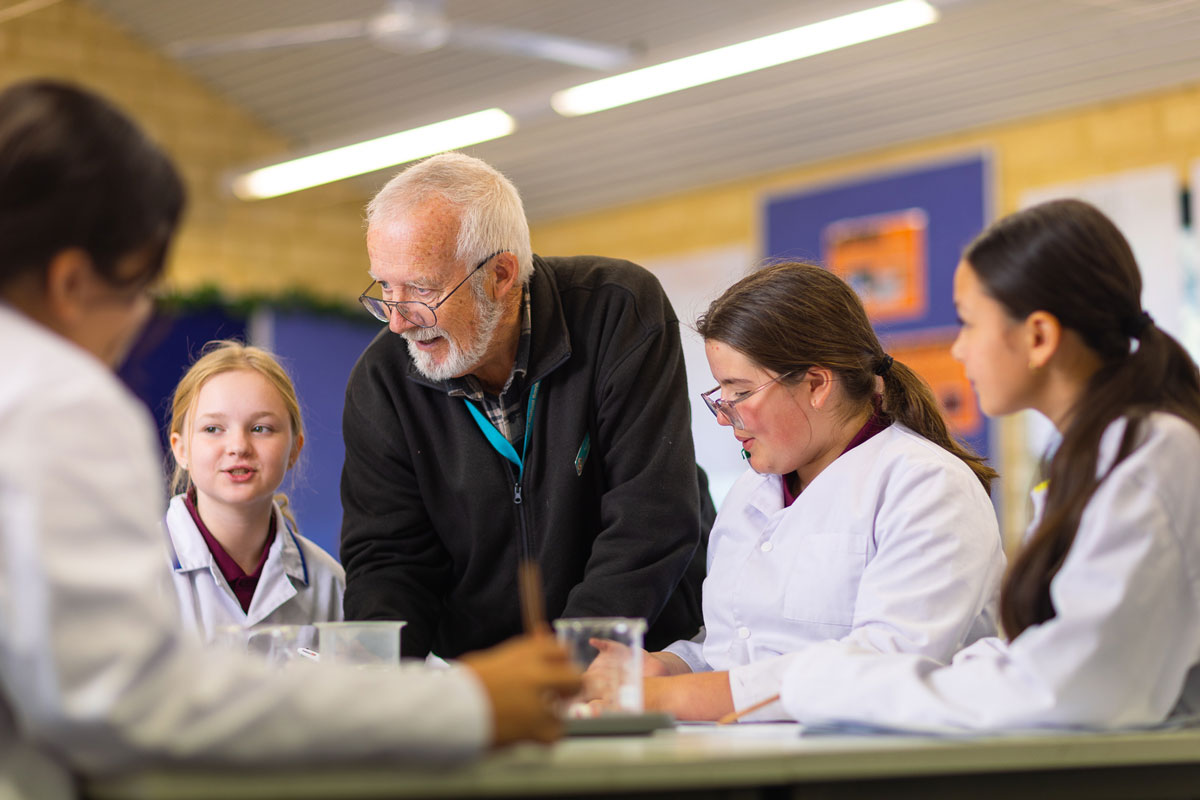 A male teacher talks to a group of female students wearing lab coats. 