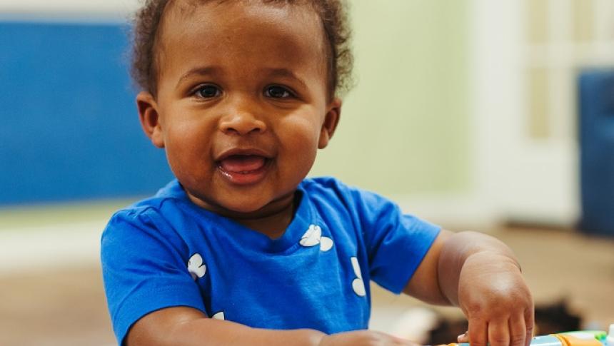 A small boy in a blue shirt in an early learning setting, looking at the camera and with his hands on a colourful toy. 