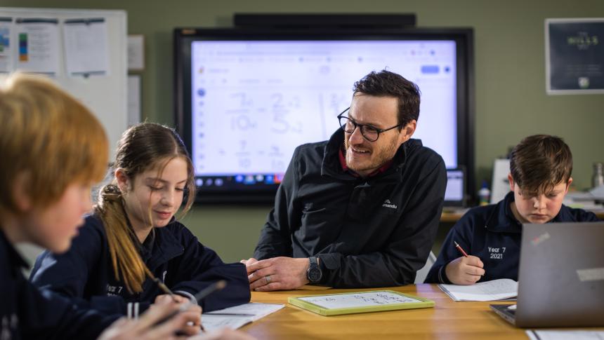 A secondary classroom setting with a male teacher sitting at a table with three students. They hold pens with paper and laptops in front of them. 