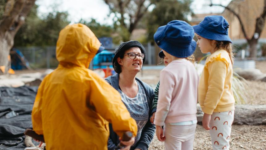 An early childhood educator outside with three children. The educator is kneeling down to speak to them at their level. 