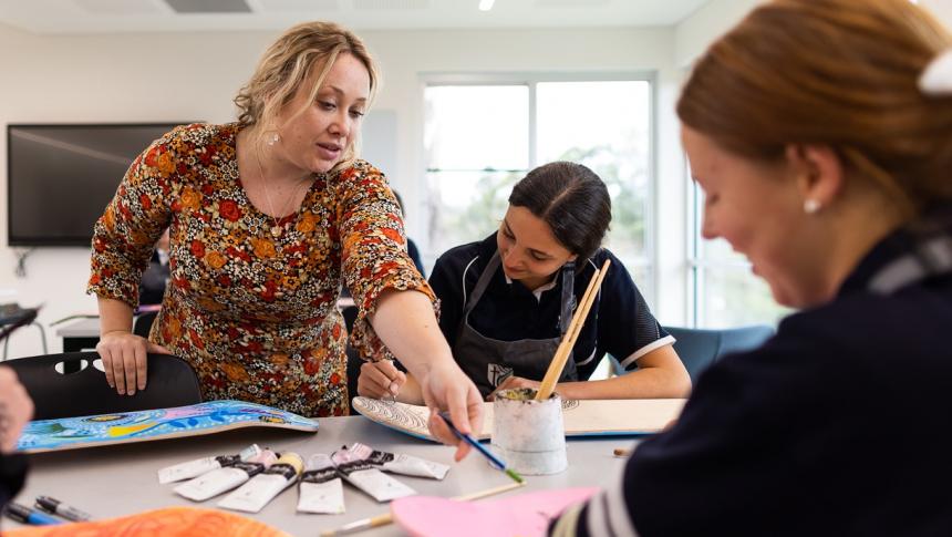 A teacher and 2 students in a secondary setting. The teacher is providing instruction and pointing at a students work. 