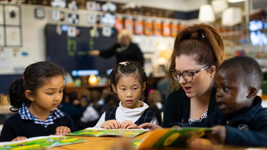 A teacher sitting at a low desk with several primary school students helping them with reading