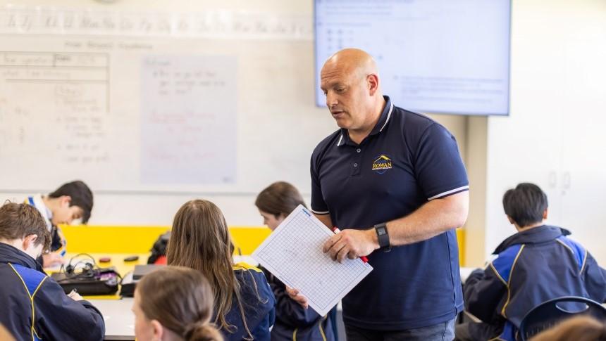 Teacher standing in the middle of his classroom supervising and assisting while students around him are working at their desks