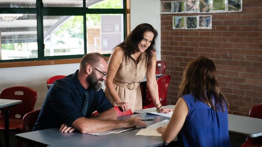 Two female and one male teacher gather around a desk in a school office discussing papers on a desk in front of them