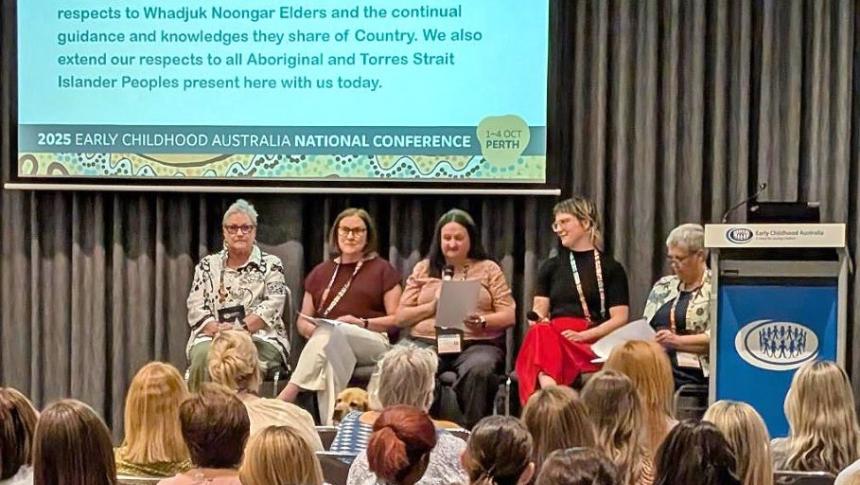 Five women sit on stage in front a screen, in the foreground an audience watch their presentation