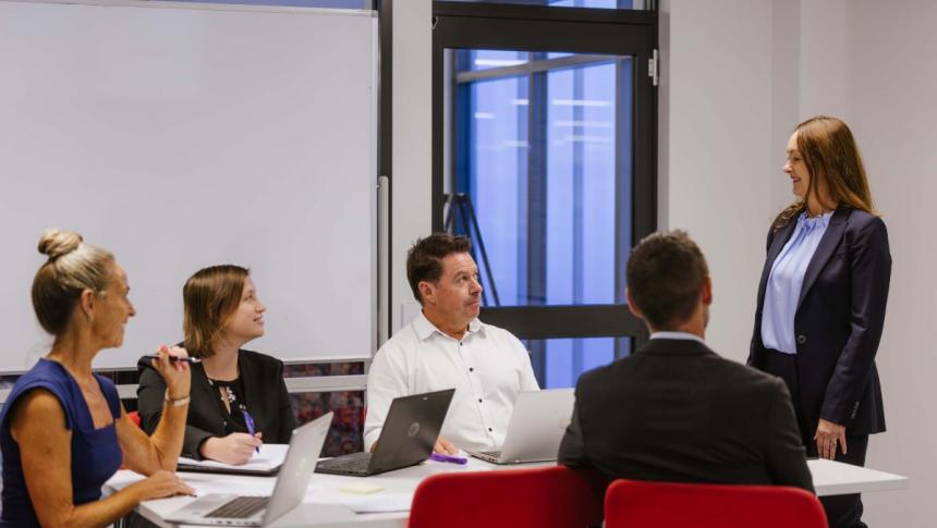 A woman stands addressing a group of teachers seated around an office desk with laptops and notebooks in front of them