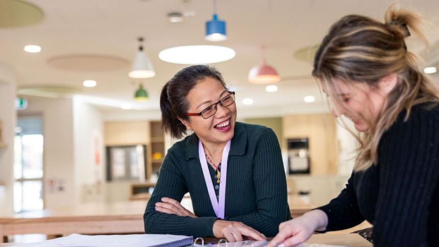 Two educators sit at a desk smiling and pointing at a folder open in front of them