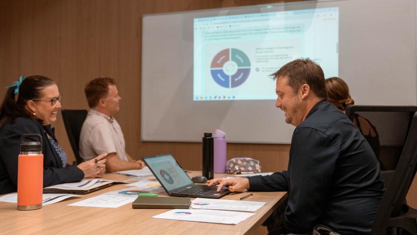 A group of professionals are seated at a desk looking at a presentation on a whiteboard