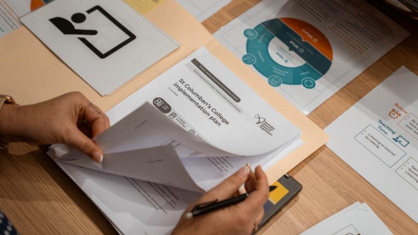 Photo of a teacher's hands as she looks through a document entitled 'St Columban's College implementation plan', at a table with other implementation plan documents