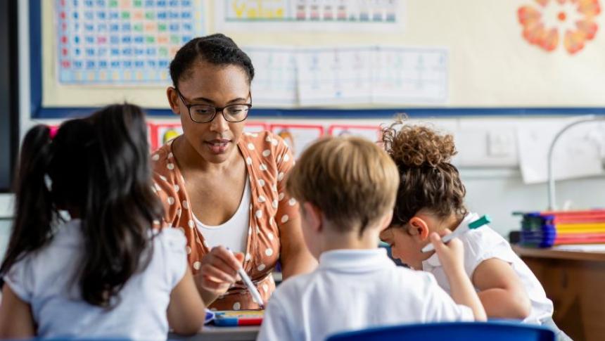 A teacher sits in front of three students with their backs to the camera, she is pointing at a piece of paper and explaining the lesson