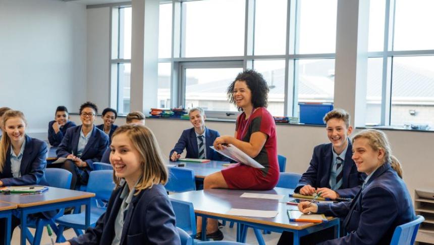A teacher sits on a desk surrounded by students, all are smiling and looking to the front of the class