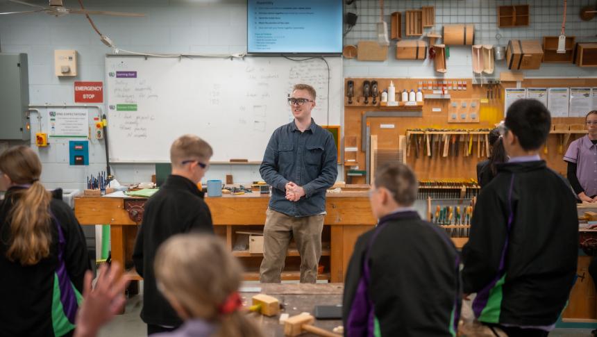 A woodworking teacher stands in front of his high school class, he is wearing safety glasses and speaking to the students