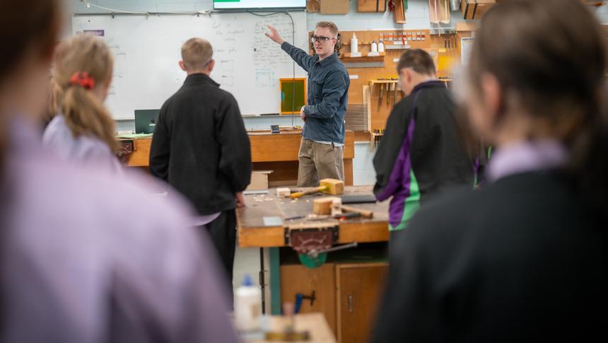 Teacher in a woodworking class in front of students, explaining text on a whiteboard.
