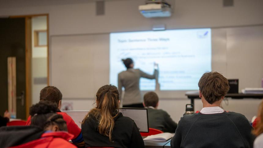 A teacher writing on a whiteboard, with a class taking notes