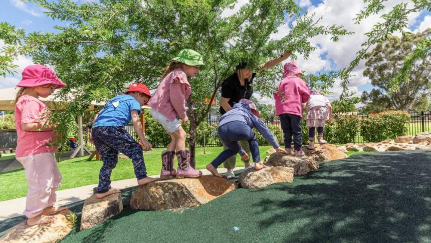 Children walk across a series of rocks in a playground under the supervision of an early childhood educator