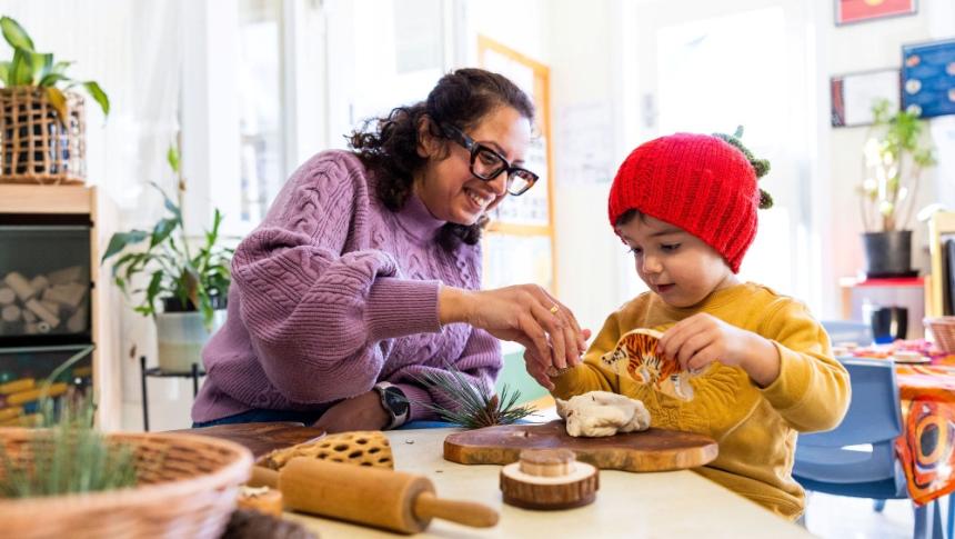 An early childhood educator with a young child in a classroom, playing with a range of materials on a wooden board