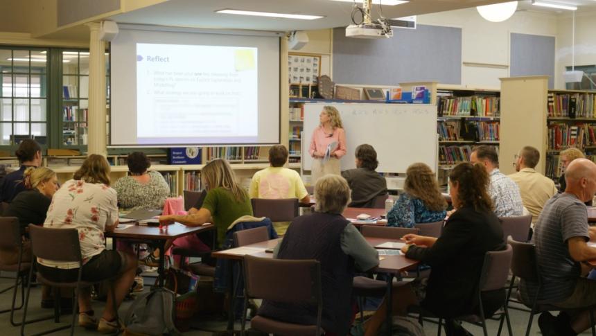 A teacher stands next to a projector screen in a library, in front of a class full of mature students