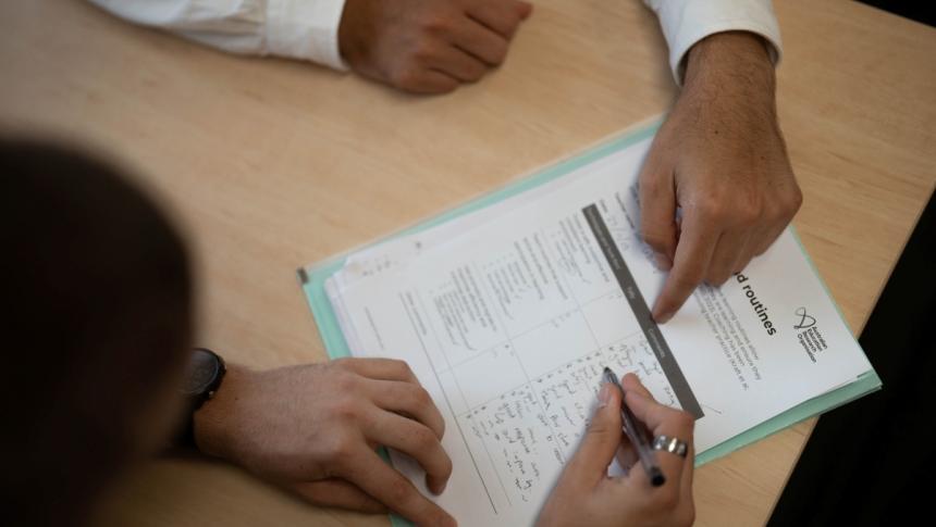 Two teachers sit at a table completing a checklist document, viewed from above with only their hands visible