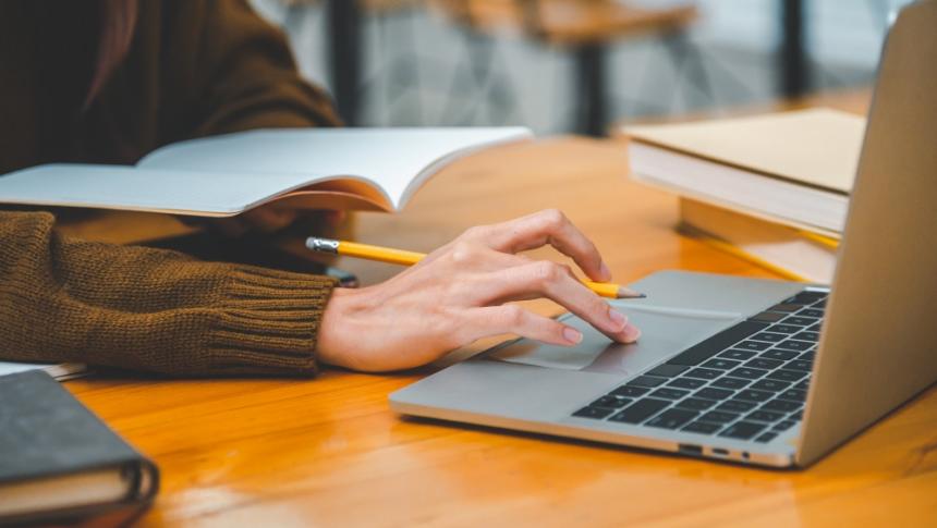 A teacher using a laptop while making notes in a notebook, only the hands are visible in frame