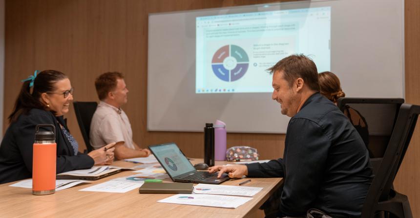 A group of professionals are seated at a desk looking at a presentation on a whiteboard