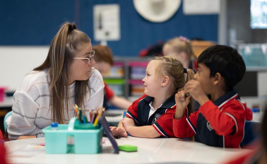 A teacher sits at a desk talking to two students