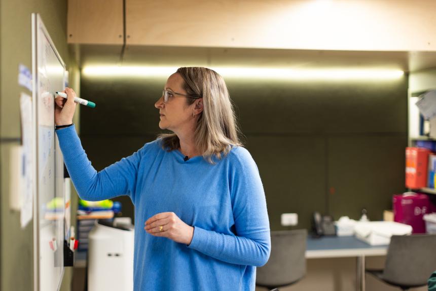 A teacher stands in front of a whiteboard, she is writing on the board with a green whiteboard marker