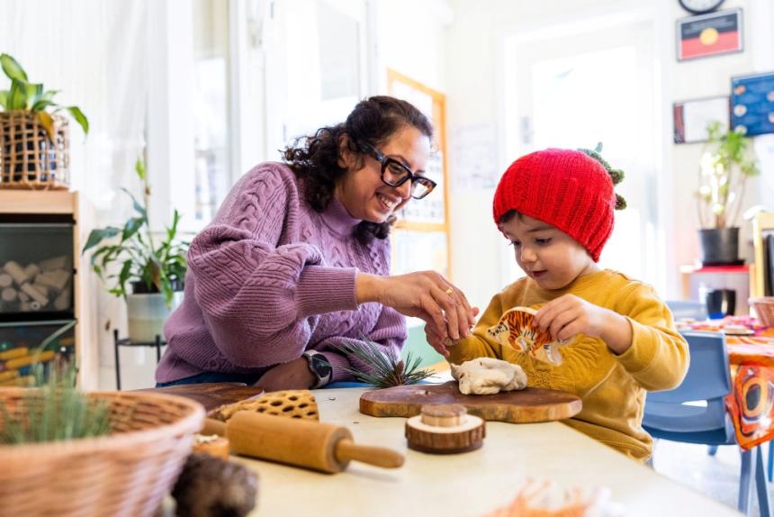 An early childhood educator with a young child in a classroom, playing with a range of materials on a wooden board